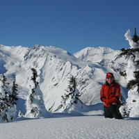A woman in a red jacket kneeling on snow in the backcountry. 