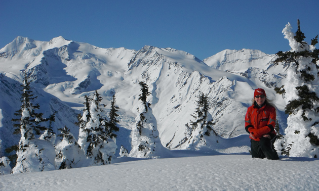 A woman in a red jacket kneeling on snow in the backcountry. 