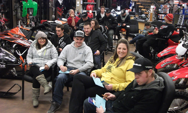 A group of people sitting on chairs in a snowmobile shop. 