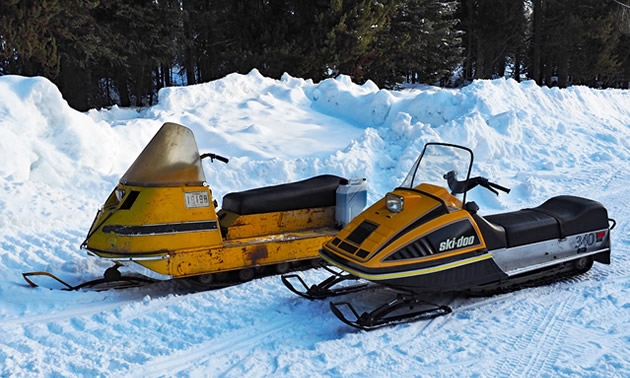 Two vintage Ski-Doo snowmobiles sit side by side on a snow covered trail 