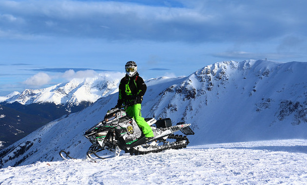 Curtis Hofsink sitting on his bright yellow sled with the mountains and blue sky in the background. 