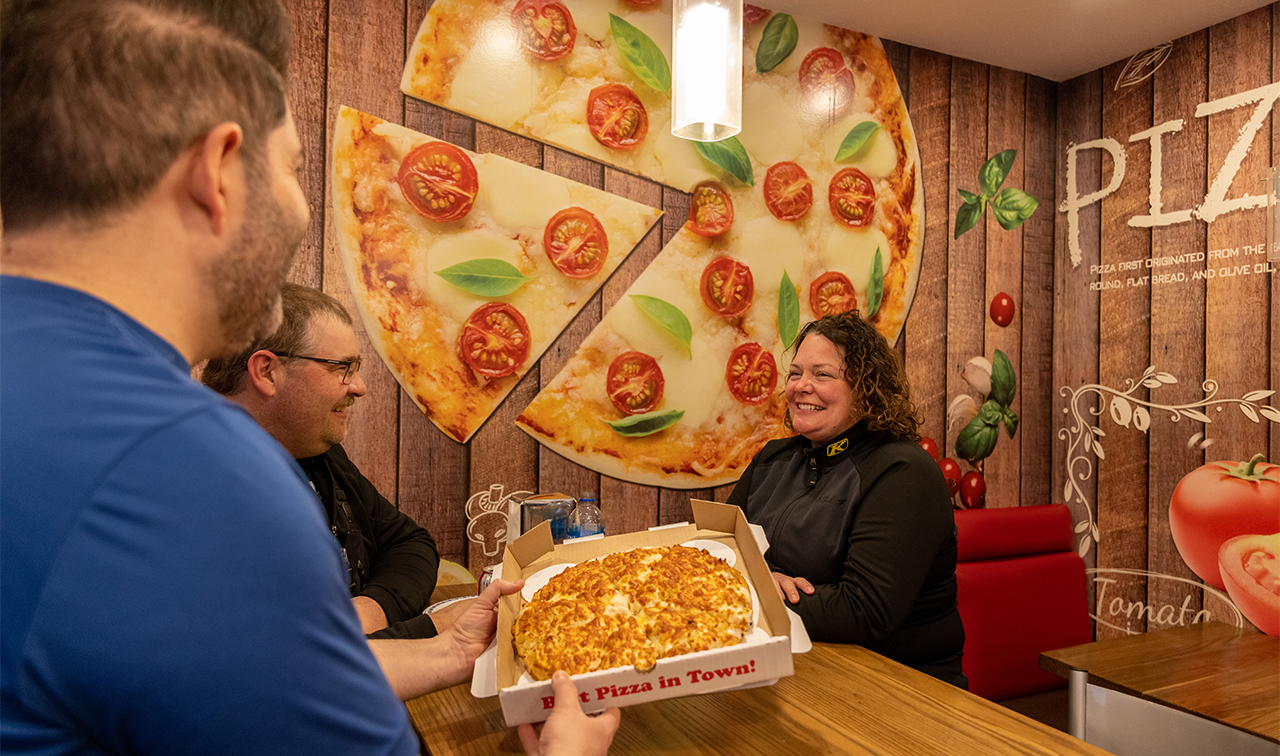 Three people sit around a table smiling at each other, about to eat pizza