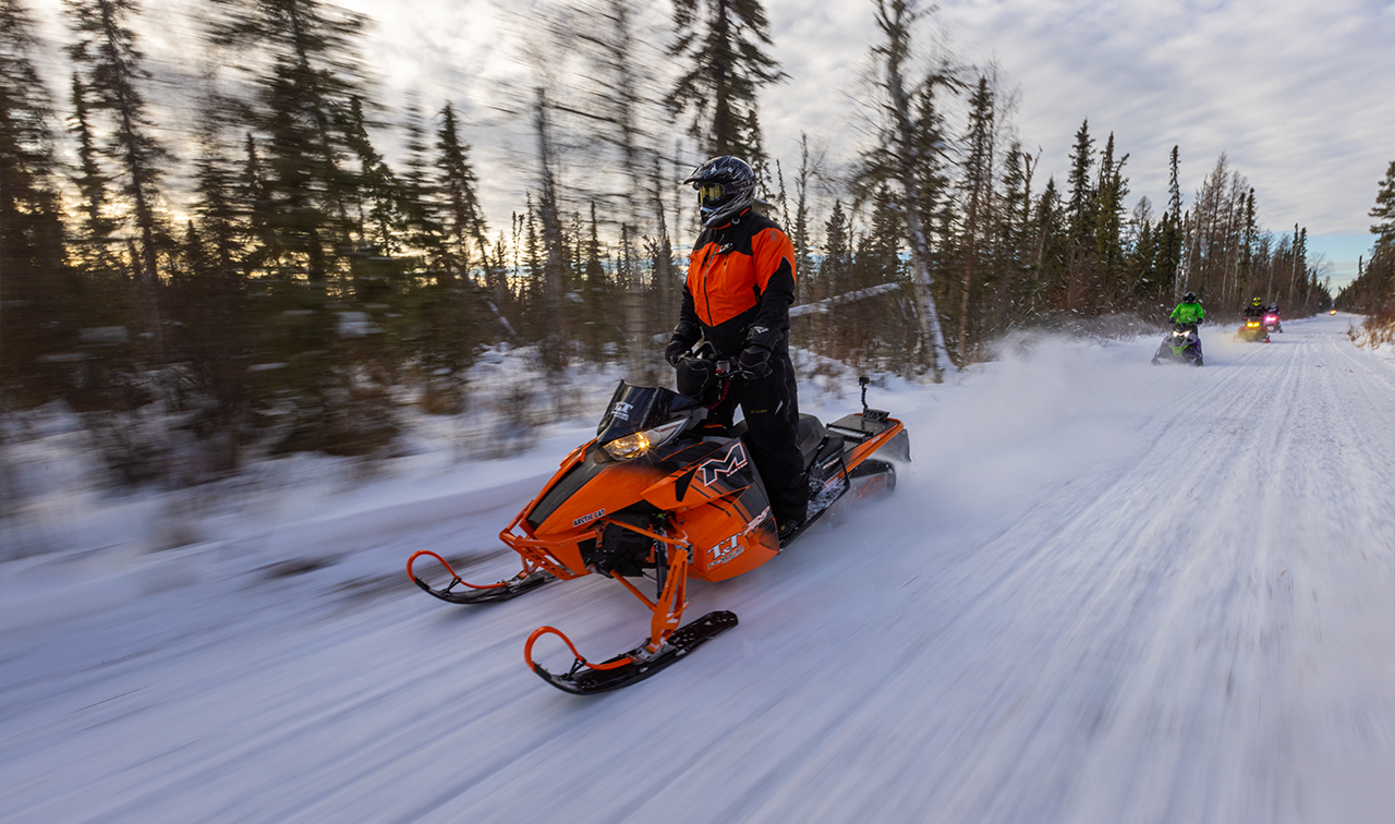A man on an orange snowmobile stands up while whooshing by on a snowmobile trail