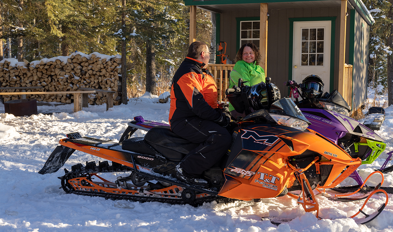 Two snowmobilers smile side-by-side next to the Legion Lodge