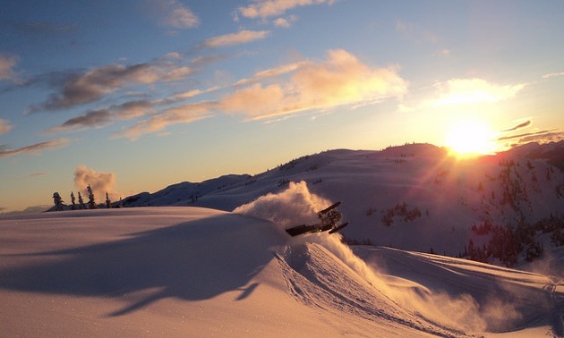 A stunning photo of the sun just setting over a mountain in the distance.  The snow looks pink in color from the reflection of the sun.