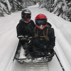 Two kids sitting in a toboggan behind a snowmobile. 