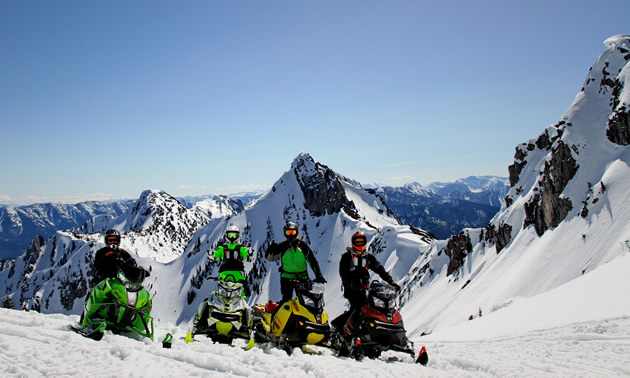 Four snowmobilers wave on a clear day in the mountains.