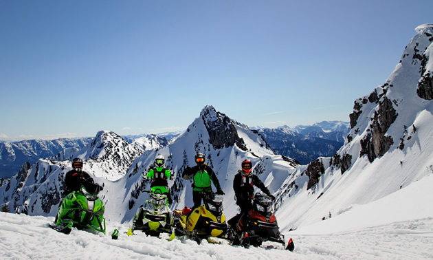 Four snowmobilers wave on a clear day in the mountains. 
