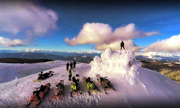 A man stands on the top of an ice sculpture igloo built on the top of a mountain with friends and their snowmobiles in front of the monument. 