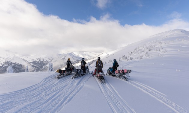 Four sledders making tracks in fresh powder and overlooking a mountain landscape
