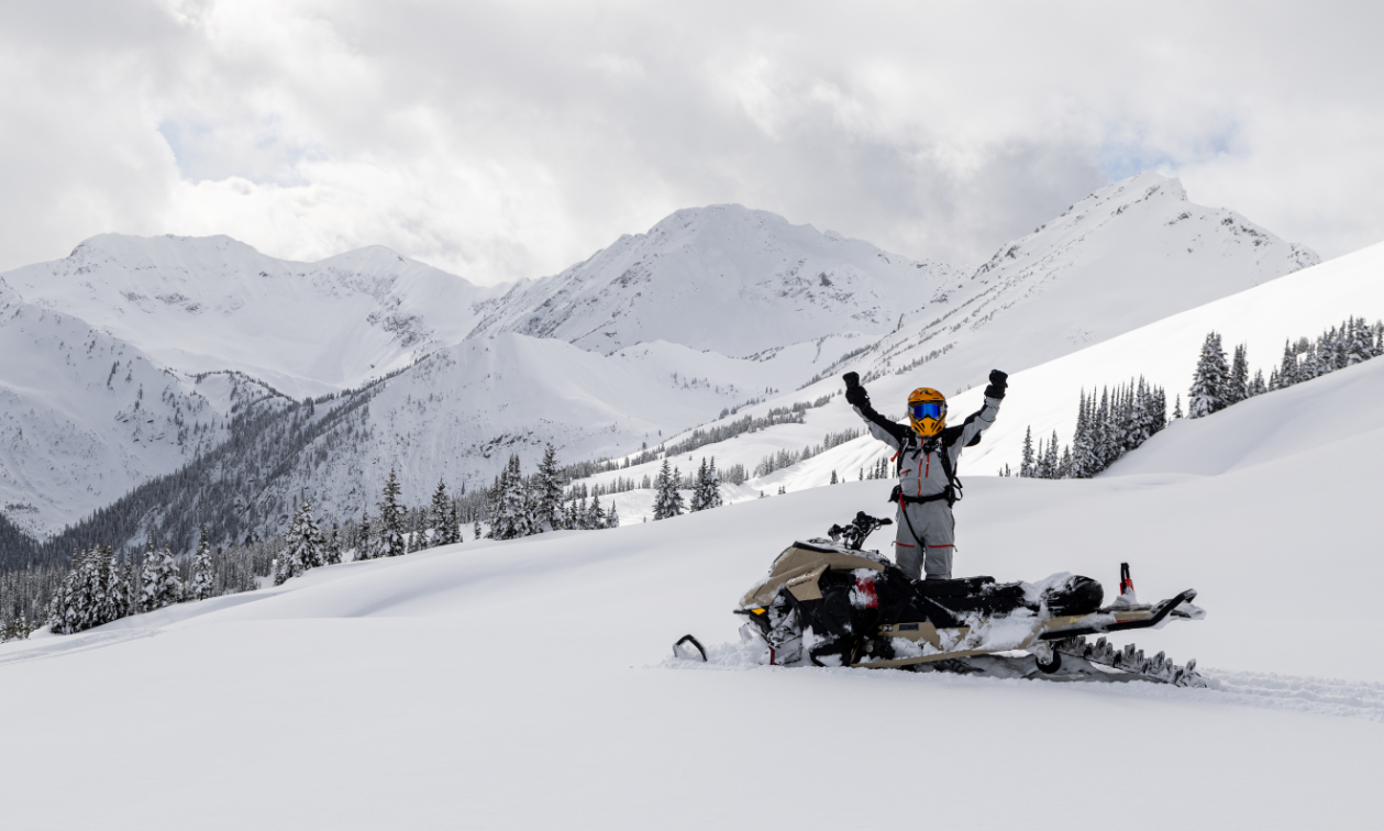 Snowmobiler standing up with arms in the air while on a snowy mountain
