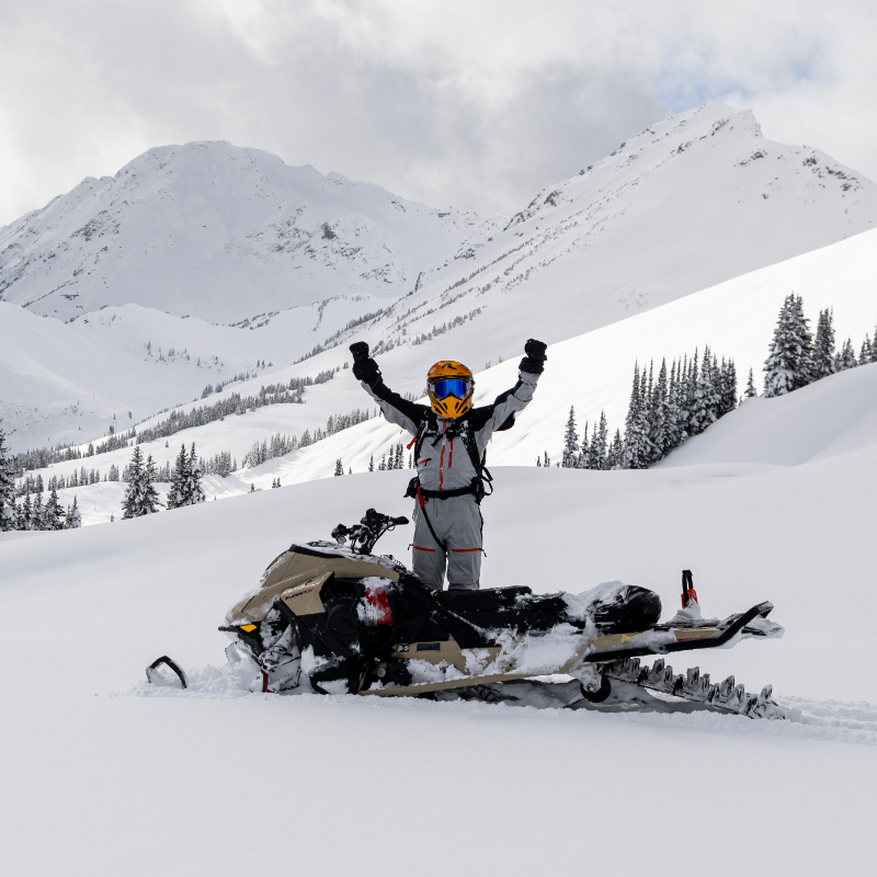 Snowmobiler standing up with arms in the air while on a snowy mountain
