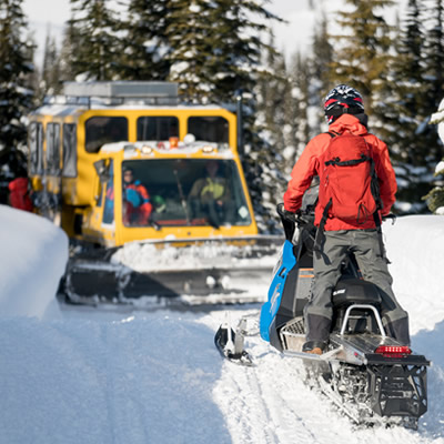 A snowmobiler is on a trail, facing a snowcat, and has no room to pass by.