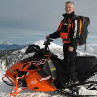 A man standing on an orange Arctic Cat in the backcountry. 
