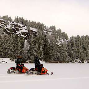 Two sleds riding in Whiteshell.  There is a rocky hill behind them and they are riding through a large open meadow.